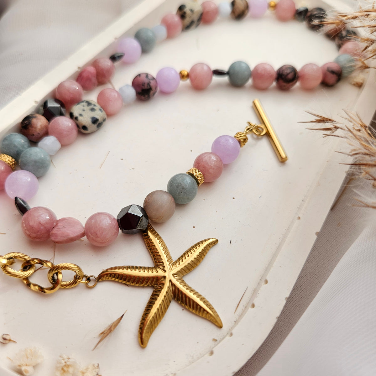 A close-up of a beaded strand jewelry piece with pastel pink, lavender, gray, and speckled dark beads, gold-toned spacer rings, a gold toggle clasp, a gold starfish charm, and a gold lobster clasp, displayed on a white surface.