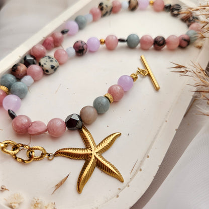 A close-up of a beaded strand jewelry piece with pastel pink, lavender, gray, and speckled dark beads, gold-toned spacer rings, a gold toggle clasp, a gold starfish charm, and a gold lobster clasp, displayed on a white surface.