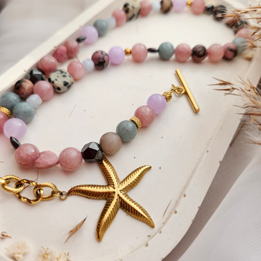 A close-up of a beaded strand jewelry piece with pastel pink, lavender, gray, and speckled dark beads, gold-toned spacer rings, a gold toggle clasp, a gold starfish charm, and a gold lobster clasp, displayed on a white surface.